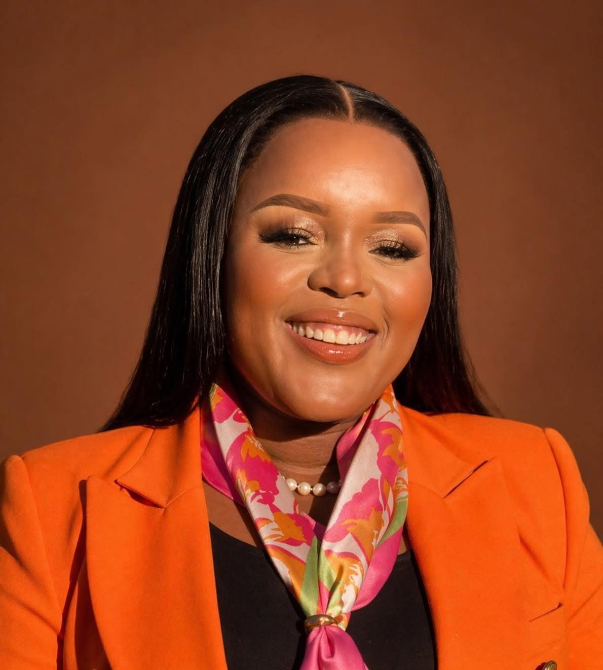 Portrait of Segolame Thaga smiling, wearing an orange blazer and silk scarf